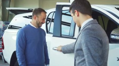 Gösterişli bir araba salonunda sakallı iyi giyimli bir satıcı el değmemiş beyaz bir aracın kapısını açar ve gülümseyen bir alıcıyı içeri davet eder. Onlar dostça bir tartışmaya girerken atmosfer heyecan ve heyecanla doludur.