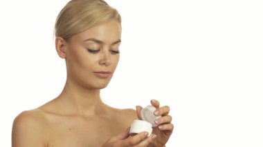 Uzun sarı saçlı ışıl ışıl genç bir kadın beyaz bir krema kavanozunu açarken parlak bir şekilde gülümsüyor. Şakacı bir şekilde parmağına sürerek, izleyiciyi el değmemiş beyaz bir arka plana karşı güzellik rutinini paylaşmaya davet ediyor.