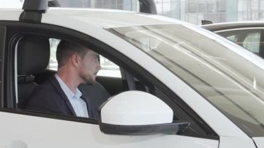 Bakımlı sakallı neşeli, yakışıklı bir adam bir galeride lüks bir arabanın içine dalmış. Onun ışıltılı gülümsemesi heyecan ve memnuniyeti yansıtıyor, konforu birleştiren modern bir otomobil seçmenin heyecanını temsil ediyor.