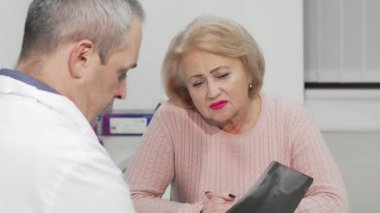 Yaşlı bir kadın doktorunun karşısında oturuyor, sağlığıyla ilgili rahatsız edici haberleri işlerken yüzünde endişeli bir ifade var. Steril klinik ortamı, teşhisin duygusal ağırlığını vurgulayarak o anın ciddiyetini vurguluyor.