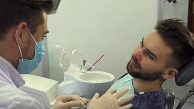 Yetenekli bir beyaz dişçi, eldiven ve maske takıyor, dişçi muayenehanesindeki genç sakallı erkek hastaya diş tedavisini tutkuyla açıklıyor. Yakışıklı esmer adam, dişçi sandalyesinde rahatça oturuyor, dikkatle dinliyor,