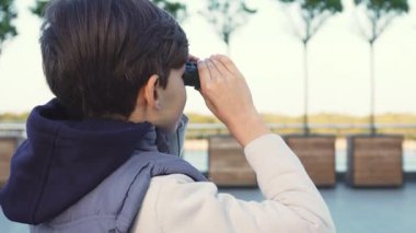 Sevimli çocuk, kolsuz beyaz gömlek ve rahat kırmızı ve yeşil kareli eşarp giymiş, dürbünle akranları, parlak bir gülümsemeyle ışıldayan yüzü. Canlı bir şehirle çevrelenmiş, macera ve merak ruhunu somutlaştırıyor.