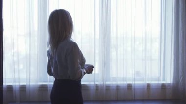 Zarif kadın panoramik bir pencerenin yanında duruyor, elinde dumanı tüten bir fincan kahveyle dışarı bakıyor, düşüncelere dalmış. Gıcır gıcır beyaz bir gömlek ve gösterişli bir etek giyerek profesyonelliği somutlaştırıyor ve rahat otel odasında güneş ışığı altında duruyor.