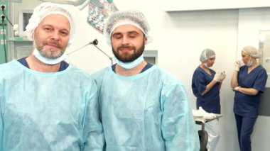 Biri sakallı ve olgun iki beyaz cerrah steril bir ameliyathanede uzmanlıklarını sergiliyorlar. Arkalarında, iki hemşire sohbete dalıp takım çalışmasından ve profesyonellikten oluşan dinamik bir atmosfer yaratıyorlar.