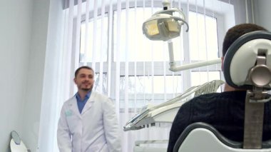 Orta yaşlı erkek bir dişçi, güvenle gülümsüyor, parlak diş lambasını hastaların ağzını aydınlatacak şekilde ayarlıyor. Kafkas uzmanı önlüğün erkeğin göğsüne mükemmel bir şekilde yerleştirildiğinden emin olmak için kapsamlı bir kontrole hazırlanıyor.