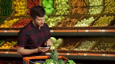 Yakışıklı, genç, beyaz bir adam akıllı telefonunu canlı taze sebze ve meyvelerin önünde dururken kullanıyor. Alışveriş arabası renkli ürünlerle dolu, hareketli bir atmosfer yaratıyor.