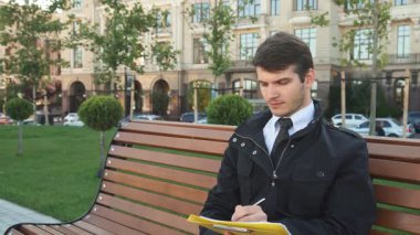 Keskin siyah takım elbiseli ve ceketli genç bir adam ahşap bir bankta, sarı bir klasöre notlar yazarak dalgın dalgın oturuyor. Yeşillik ve şehir mimarisi ile çevrelenmiş bir sonraki büyük hamlesini planlarken azim ve profesyonelliği somutlaştırıyor.