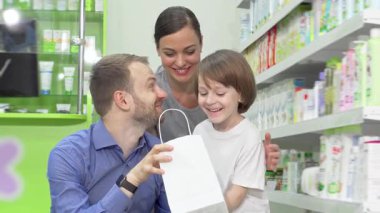 Gülümseyen bir baba, anne ve onların sevimli oğullarından oluşan neşeli bir aile, şık bir alışveriş çantasından aldıkları eczaneyi hevesle inceliyor. Sağlık ürünleriyle dolu renkli raflarla çevrili bu iç açıcı sahne,