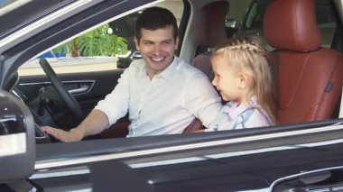 Gösterişli, modern bir arabada, neşeli bir baba, küçük kızını hareketlerle meşgul eder, çeşitli düğmelere ve özelliklere işaret eder. Büyüleyici bir menekşe elbise giymiş olan kız, dikkatle dinlerken neşe dolu ışınlar saçarak, iç açıcı bir...