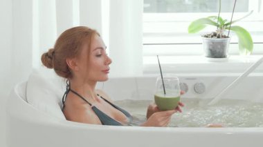 Büyüleyici bir kadın, rahatlatıcı su ve yumuşak ışıkla çevrili lüks bir girdap banyosunda dinlenirken ferahlatıcı bir detoks smoothie içiyor. Bu dingin spa anı refahın ve gençleşmenin özünü yakalar, sizi kendini şımartmaya davet eder.