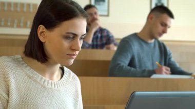 Nazik bir beyaz kız öğrenci, düşünceli bir ifadeyle, geniş bir konferans salonunda otururken dizüstü bilgisayarına notlar yazar. Arka planda, iki erkek sınıf arkadaşı derslerine devam ediyor, biri deftere yazıyor, diğeri ise derin.