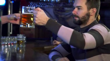 Yakışıklı, sakallı bir adam bardaki bir bardak biranın üzerine uzanıp zengin aromanın tadını çıkarıyor. Barın sıcak atmosferi, kırsal dekoru ve yumuşak ışıklandırmasıyla, bir yudum almaya hazırlanırken,