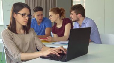 Kendine güvenen esmer bir kız, parlak, modern bir ofis masasında dizüstü bilgisayarında yazı yazarken başparmağını kaldırıyor. Arka planda, üç iş arkadaşı canlı bir tartışmaya girişerek takım çalışmasının ve yaratıcılığın dinamik atmosferini yaratıyorlar.