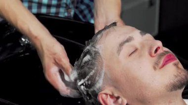 Profesyonel bir kuaför saçlarını nazikçe yıkar ve berber dükkanında sakin bir atmosfer yaratır. Rahatlatıcı su ve bol köpük başını sarar, ferahlatıcı bir saç kesimi deneyimi vaad eder.