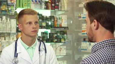 Odaklanmış esmer bir adam, beyaz önlük ve steteskop giyerken dikkatle dinleyen genç bir doktorla sohbet eder. Çeşitli ilaçlarla dolu renkli raflarla çevrili bu sahne sağlık hizmetlerinin özünü yakalıyor.