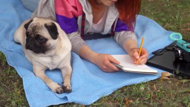 Genç bir kız, güneşli bir parkta parlak mavi bir battaniyenin üzerinde rahatlar ve dikkatle defterine yazar. Çekici köpeği yanında uzanıyor, kameraya şen şakrak bakıyor, doğadaki dostluk ve rahatlığın özünü yakalıyor.