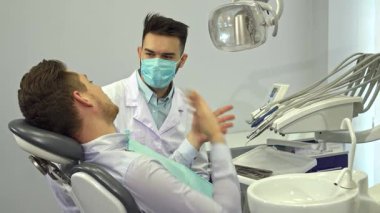 Şık sarı saçlı neşeli genç bir adam modern bir dişçi koltuğunda otururken başparmağını kaldırıyor, kendine güven ve memnuniyet saçıyor. Dost canlısı diş uzmanı, maske ve eldiven takmış, onun yanında duruyor ve onu karşılıyor.