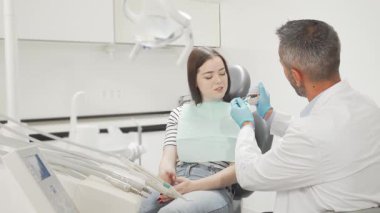 Neşeli genç bir kadın modern bir dişçi koltuğunda oturmuş diş kalıbını gösteren dişçiyle meşgul oluyor. Parlak, steril çevre güven ve profesyonelliği yayar, diş bakımını ulaşılabilir ve bilgilendirici kılar.