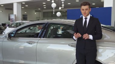 Karizmatik bir satış müdürü, modern bir bayilikte çarpıcı yeni gri bir sedanın yanında parlak bir gülümseme ile parlak bir araba anahtarı sergiliyor. Sivri siyah takım elbiseli ve kravatlı bu genç beyaz adam profesyonelliğini sergiliyor.