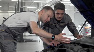 Gri üniformalı iki yetenekli oto tamircisi kaputu açık bir arabanın üzerine eğilip motor sorununu teşhis etmeye çalışıyor. Odaklanmış ifadeleri ve işbirliği çabaları otomotiv tamir sektöründe gerekli takım çalışmasını vurguluyor