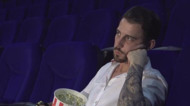 Loş bir tiyatroda, koyu saçlı ve dövmeli genç bir adam koltuğunda oturur, gözlerini açık tutmak için çabalarken açıkça sıkılır. Elinde yarısı yenmiş patlamış mısırla, sönük bir film boyunca uyanık kalma mücadelesini somutlaştırıyor.