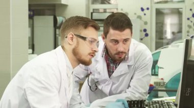 Beyaz laboratuvar önlükleri ve lateks eldivenleri giymiş iki özenli erkek laboratuvar asistanı, bir bilgisayara yaslanmış, ekranda gösterilen test sonuçlarını tartışmakla meşguller. İfadeleri işbirliği yaparken merak ve kararlılığı yansıtır.