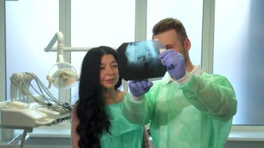 Yeşil üniformalı, odaklanmış sarışın bir erkek dişçi yaşlı bir esmer kadın hastaya x-ray bulgularını tutkuyla açıklıyor. Sahne, dişçinin x-ray görüntüsünü göstererek profesyonel bir diş ortamını görüntüler.