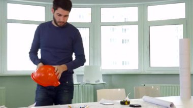 Kendini işine adamış genç esmer bir mimar masaya eğilip parlak bir ofisin planlarının yanına turuncu bir şapka takıyor. Ciddi bir beyaz erkek inşaat mühendisi, bakımlı sakallı, arka planda güvenle duruyor.