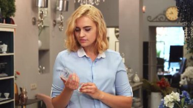 Şık bir ev dekorasyonu mağazasında güzel sarı kıvırcık saçlı genç bir kadın incelikle zarif gözlükler seçiyor. Her parçayı gülümseyerek inceler, kadehlerin kusursuz ve rahat yaşam alanı için mükemmel olduğundan emin olur.