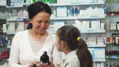 Renkli ürünlerle dolu parlak bir eczanede gülümseyen bir anne, ilacı birlikte incelerken meraklı kızıyla bir anı paylaşıyor. Küçük kız, uzun siyah saçları arkadan bağlanmış, heyecanla yukarı bakıyor, seçimlerden büyülenmiş.