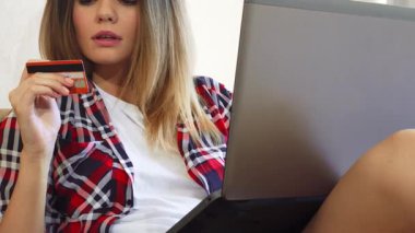 Genç bir kadın rahat bir şekilde rahat odasında oturuyor ve online alışverişler yaparken dizüstü bilgisayarına odaklanıyor. Tatmin olmuş bir gülümseme ile, kredi kartını tutuyor, şık bir kareli gömlekle evden alışveriş yapmanın neşesini somutlaştırıyor.