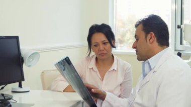 Parlak bir doktor ofisinde genç bir kadın, doktorunun röntgen sonuçlarını dikkatlice dinlerken gülümsüyor. Sıcak atmosfer, bej renkli yumuşak duvarlar ve beyaz giyinmiş doktorun profesyonel tavırlarıyla güçlenir.