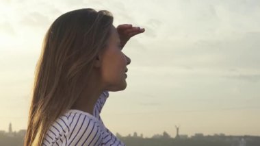 Neşeli genç bir kadın, nefes kesen şehir manzarasına hayranlıkla bakarken, gözlerini güneşten koruyor. Uzun, siyah saçları akşam rüzgarında hafifçe sallanırken kameraya doğru döner ve parıldayan bir gülümseme yakalar.