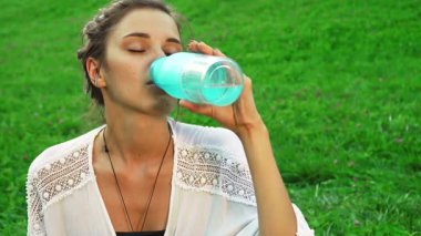Genç bir kız dinlendirici bir yudum su almak için yoga pratiğini durdurur, huzur ve mutluluk yayar. Parlak bir gülümseme ve iyi niyetli bir jest ile yemyeşil bir parkın ortasında sağlıklı bir yaşam tarzının özünü temsil ediyor.