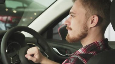 Yeni arabasının şoför koltuğunda oturan neşeli, sakallı, genç bir adamın yakın çekimi. Araba anahtarlarını güvenle sergiliyor. Parlak gülümsemesi sahipliğin heyecanını ve yeni maceralara atılmanın heyecanını yansıtıyor.