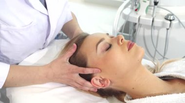 Gıcır gıcır beyaz önlüklü yetenekli bir kozmetikçi müşterinin kafasına nazikçe masaj yapıyor, kulaklarına ve şakaklarına odaklanıyor. Güzellik salonunun sakin atmosferi deneyimi artırıyor. Güzel sarışın kadın, akubasıncını tazelemekten hoşlanıyor.