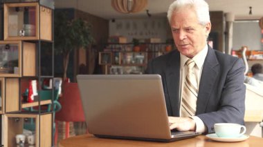 Sivri bir takım elbise ve kravat takmış seçkin bir Kafkas işadamı, bir fincan kahvenin tadını çıkarırken laptopuna dikkatle odaklanıyor. Modern dekor ve sıcak aydınlatmayla süslenmiş rahat kafe dekoru, davetkar bir atmosfer yaratıyor.