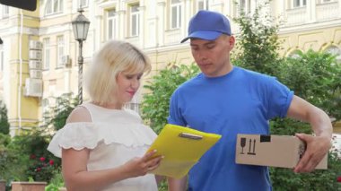 Havalı beyaz bluzlu mutlu bir kadın, mavi şapkalı yakışıklı bir kuryeden karton bir paket alırken fiş imzalar. Bu canlı açık hava sahnesi çevrimiçi alışverişin heyecanını yakalıyor.