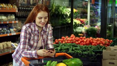 Şık, kızıl saçlı genç bir kadın akıllı telefonuyla alışveriş listesini karıştırırken gülümsüyor, market arabasına yaslanıyor ve içi taze sebze ve meyve dolu. Süpermarketin renkli arka planı, canlı bir şekilde süslenmiş.