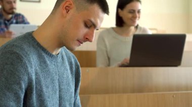 Kafkasyalı genç bir erkek öğrencinin yakın çekimi, bilimsel bir kitaba çok dalmış, bir konferans salonunda konsantrasyonunu sergiliyor. Arka planda, bir kadın dizüstü bilgisayarında yazarken, başka bir erkek tabletle meşgul olarak dinamik bir akademik kurgu yaratıyor.