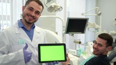 Dost canlısı, orta yaşlı beyaz bir dişçi, canlı yeşil krom bir ekranla dijital tableti gururla sergiliyor. Parlak, modern bir diş muayenehanesinde dururken, gülümseyen erkek hastası...