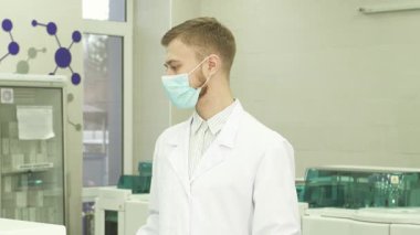 Gelişmiş ekipmanlarla dolu parlak, modern bir laboratuvarda beyaz önlüklü ve maskeli genç bir laboratuvar asistanı profesyonellik ve güven dolu başını kameraya doğru çeviriyor. Sıcak gülüşü ve özenli bakışları izleyicileri davet ediyor.