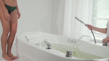 Işıl ışıl gülümseyen neşeli bir kadın, rahatlatıcı bir hidromasaj yapmaya hazır, lüks bir girdap banyosuna adım atıyor. Yumuşak doğal ışığın pencereden süzüldüğü sakin spa ortamı, rahatlama atmosferini arttırır.