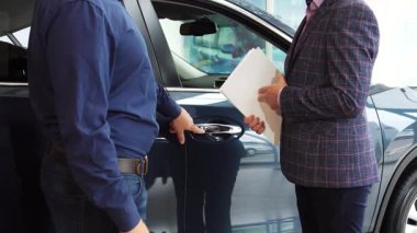 Gösterişli bir araba galerisinde, yeni sahibi yeni satın aldığı aracın karmaşık kilitlerini hevesle öğreniyor. İki iyi giyimli adam, biri mavi gömlekli, diğeri şık ceketli, sıkı bir tokalaşma paylaşıyor, başarılı bir anlaşmayı simgeliyor ve...