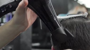 Odaklanmış bir berber, erkek müşterisinin saçını şekillendirmek için bir saç kurutma makinesini ustalıkla kullanır ve modern bir berber dükkanında cilalı bir görünüm yaratır. Sahne, profesyonel tımarın özünü yakalıyor, saç bakımındaki ustalığı ve hassasiyeti gözler önüne seriyor.