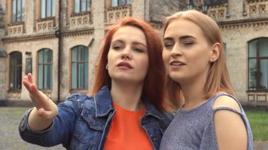 Kızıl saçlı bir kız heyecanlı bir şekilde kameranın dışındaki bir şeyi işaret ederken sarışın arkadaşı kahkahaya boğuluyor ve canlı bir atmosfer yaratıyor. Üniversiteye yakın bir yerde dururlarken, ifadeleri mutluluk ve dostluk yayar,