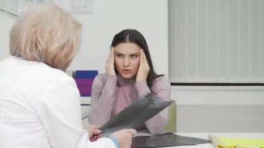 Acı içinde başını tutan sıkıntılı genç bir kadın, parlak ve modern bir ofiste doktoruna danışıyor. Kadın hasta, gözle görülür bir şekilde endişeli, şiddetli migreni hakkında tartışırken doktor dikkatli bir şekilde tıbbi taramalarını gözden geçirir, şefkat ve...