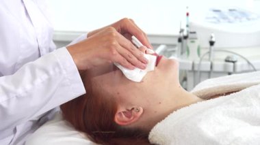 Yetenekli bir kozmetikçi kadın müşterilerinin yüzünü nazikçe yumuşak peçetelerle siler ve beyaz güzel kızı cilt tedavisine hazırlar. Dingin atmosfer, arka plandaki klinik ekipmanlarla gelişir.