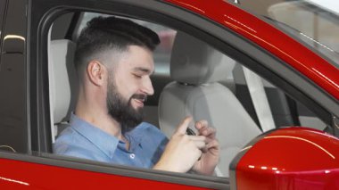 Şık sakallı neşeli bir adam çarpıcı kırmızı arabasının anahtarlarını tutarken heyecan ve gurur saçıyor. Bu canlı sahne, sahipliğin heyecanını ve otomobiller dünyasında yeni başlangıçların neşesini yakalar.