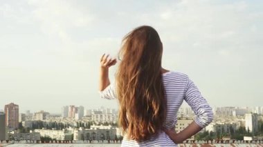 Genç bir kadın kameraya arkası dönük duruyor, uzun siyah saçları hafif meltemde dans ediyor aşağıdaki çarpıcı şehir manzarasına bakıyor. Akşam gökyüzü ufuk çizgisinde sıcak bir parıltı yayıyor, canlı binaları ve yemyeşil alanları aydınlatıyor.